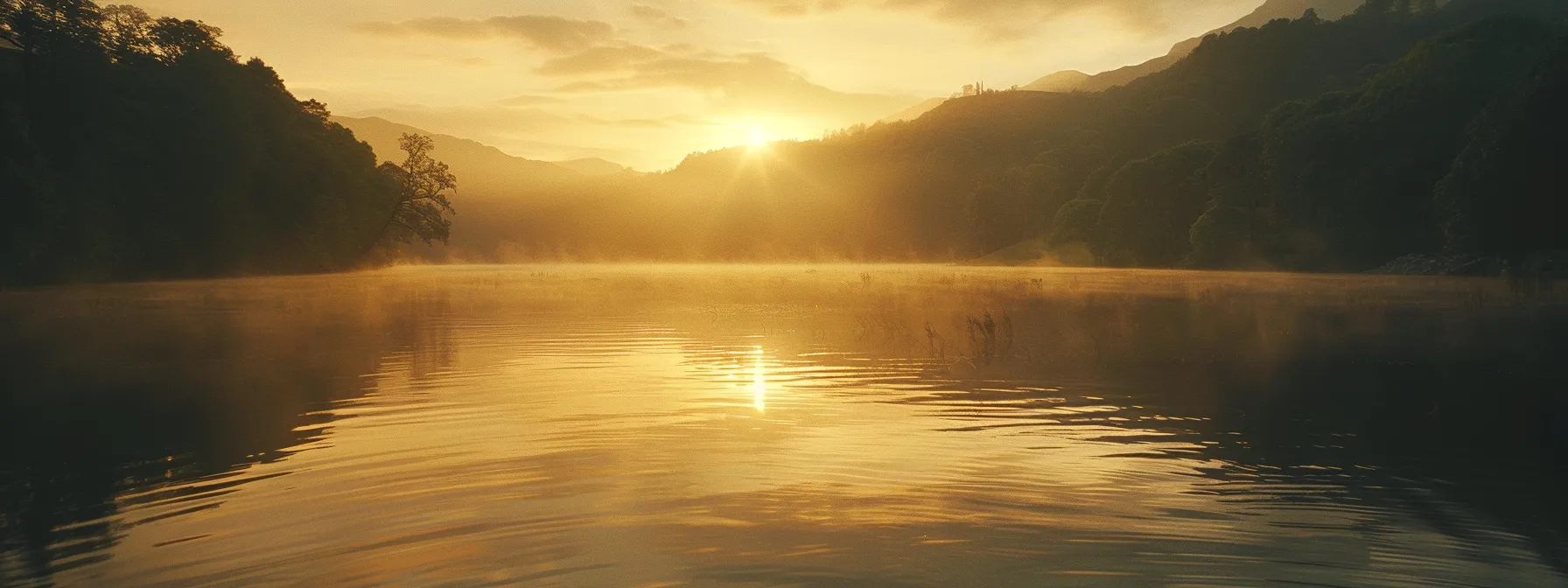 a majestic sunrise illuminates a tranquil lake surrounded by mist-covered mountains, casting a golden glow on the glassy water surface.