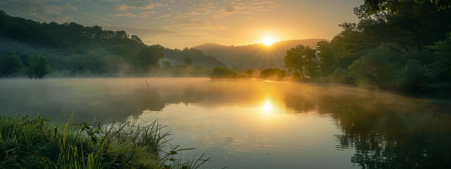 a radiant sunrise casts golden hues over a tranquil lake, where mist rises gently from the water's surface, enveloping surrounding lush green hills in a serene glow.