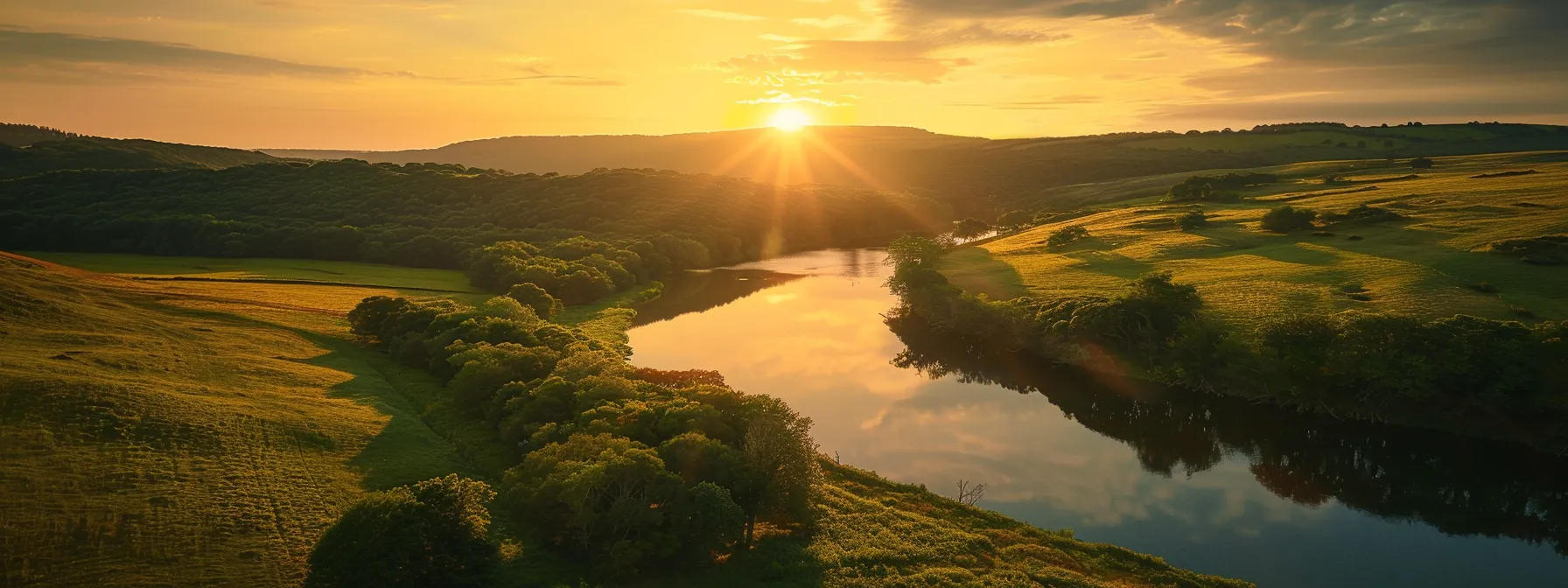 a vibrant sunset casts a warm golden glow over a serene landscape of rolling hills, accentuated by lush green trees and a tranquil river reflecting the colorful sky.