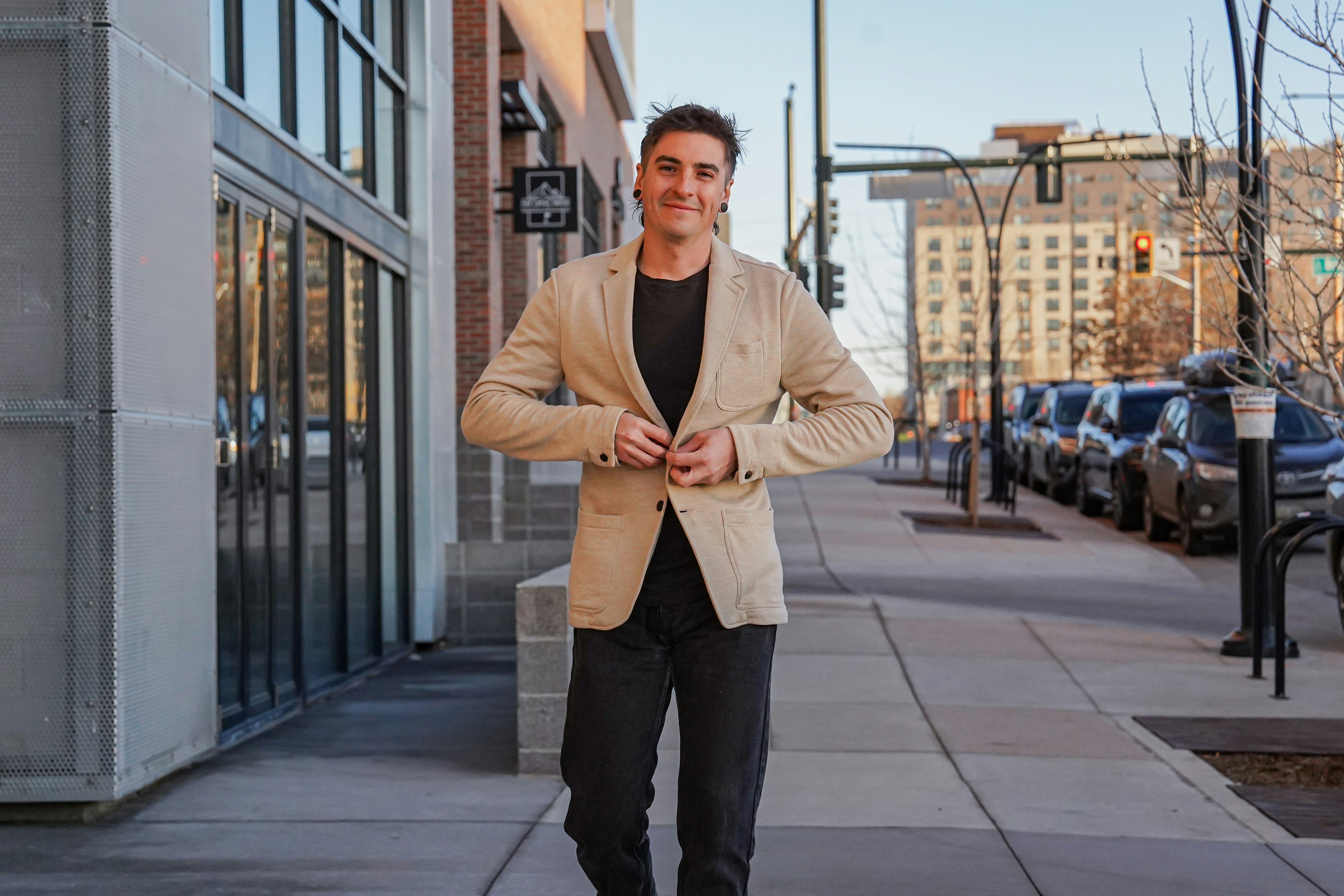 A confident young man walks down a modern city sidewalk, smiling slightly while buttoning a beige blazer, exuding casual professionalism.